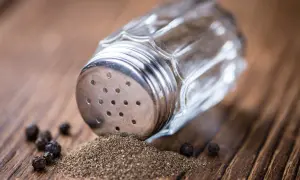 Ein Glasstreuer mit Metallaufsatz liegt auf einem Holztisch. Gemahlener Pfeffer und ganze Körner sind daneben verstreut. So kann man den Pfeffer neutralisieren.
