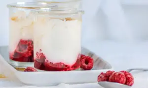 Zwei Gläser mit cremigem Dessert und Himbeeren. Unten leuchtet ein intensiver Himbeercoulis, gekrönt von luftiger Sahne. Frische Himbeeren zieren die Gläser und eine Löffelspitze.