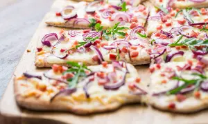 Flammkuchen ohne Speck, belegt mit roten Zwiebeln, Käse und Rucola, auf einem Holzbrett.