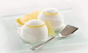 Zitroneneis selber machen: Zwei Kugeln erfrischendes Zitroneneis mit Zitronenspalten und einem Eisportionierer auf einem Glas-Tablett.