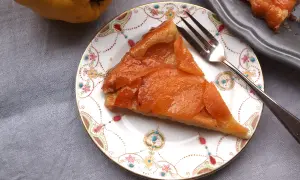 Ein Stück Quitten Tarte Tatin auf einem Teller mit Blumenmuster. Daneben eine ganze Quitte und ein weiteres Stück Tarte.