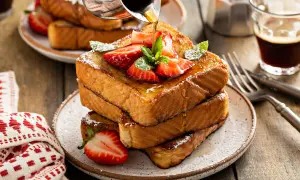 Ein Stapel goldbrauner Pain Perdu, garniert mit Erdbeeren und Minze, wird mit Sirup übergossen. Daneben ein Glas Kaffee.