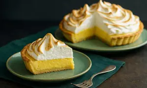 Ein Stück Tarte au Citron mit gebackener Baiserhaube auf einem Teller, daneben eine ganze Tarte.