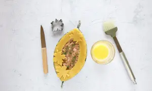 Ein ovaler Teigmantel, gefüllt mit Hackfleisch und Kräutern, wird mit Blätter-Ausstechern verziert. Daneben liegen ein Messer, ein Ausstecher, Eigelb und ein Pinsel für das perfekte Pate en croute.
