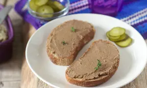 Zwei Scheiben Brot mit selbstgemachter Leberwurst, garniert mit Kräutern, dazu Gewürzgurken und ein lila Glas.