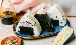 Drei dreieckige Onigiri mit Thunfisch-Mayo-Füllung, umhüllt von Nori-Algen, auf einem blauen Teller. Daneben eine Flasche Sojasauce und eine Schale mit Gewürzen.