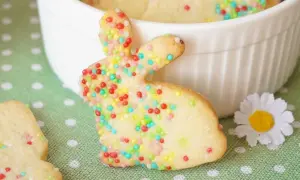Bunte Osterkekse zum Ausstechen in Hasenform, verziert mit bunten Streuseln, liegen auf einem grünen Tupfentuch neben einer weißen Schüssel.