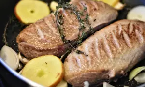 Zwei gebratene Entenbrüste mit Rosmarin, Apfelscheiben und Zwiebeln in einer Pfanne. Ein köstliches entenfilet rezept.