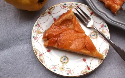 Ein Stück Quitten Tarte Tatin auf einem Teller mit Blumenmuster. Daneben eine ganze Quitte und ein weiteres Stück Tarte.