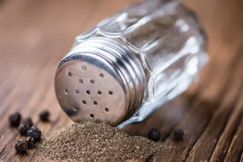 Ein Glasstreuer mit Metallaufsatz liegt auf einem Holztisch. Gemahlener Pfeffer und ganze Körner sind daneben verstreut. So kann man den Pfeffer neutralisieren.