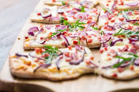 Flammkuchen ohne Speck, belegt mit roten Zwiebeln, Käse und Rucola, auf einem Holzbrett.