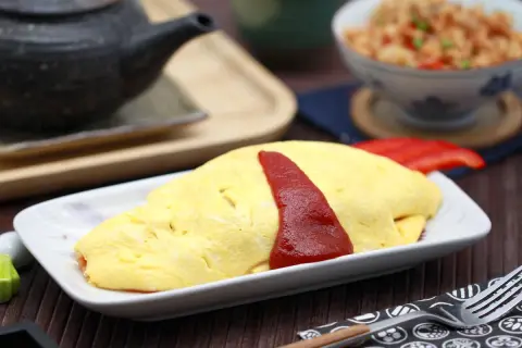 Ein leckeres Omurice Rezept: goldgelbes Omelett mit Ketchup-Streifen auf einem Teller, daneben eine Schüssel mit gebratenem Reis.