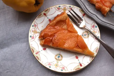 Ein Stück Quitten Tarte Tatin auf einem Teller mit Blumenmuster. Daneben eine ganze Quitte und ein weiteres Stück Tarte.