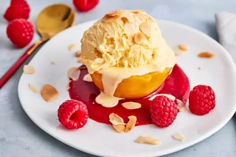 Ein Pfirsich Melba Rezept Original: Ein Pfirsich mit Vanilleeis, Himbeersauce und Mandelsplittern auf einem Teller, garniert mit frischen Himbeeren.
