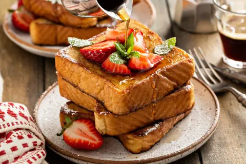 Ein Stapel goldbrauner Pain Perdu, garniert mit Erdbeeren und Minze, wird mit Sirup übergossen. Daneben ein Glas Kaffee.