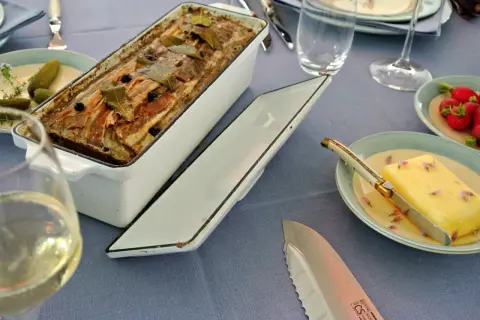Fertige Terrine mit Speck und Lorbeerblättern, bereit für ein köstliches Terrinen Rezept. Dazu gibt es Butter, Radieschen und ein Glas Weißwein.