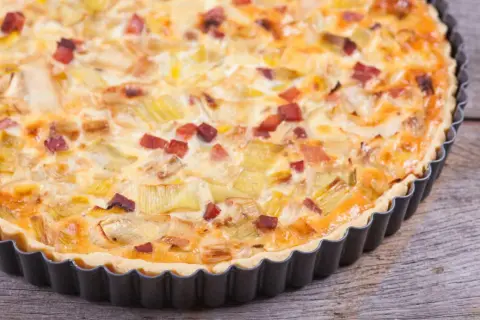 Eine herzhafte Quiche Lorraine mit Lauch und Speck in einer Metallform auf Holzhintergrund.