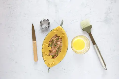 Ein ovaler Teigmantel, gefüllt mit Hackfleisch und Kräutern, wird mit Blätter-Ausstechern verziert. Daneben liegen ein Messer, ein Ausstecher, Eigelb und ein Pinsel für das perfekte Pate en croute.