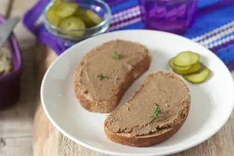 Zwei Scheiben Brot mit selbstgemachter Leberwurst, garniert mit Kräutern, dazu Gewürzgurken und ein lila Glas.