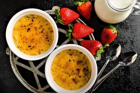 Zwei cremige Crème brûlée mit karamellisierter Zuckerkruste, garniert mit frischen Erdbeeren und einem Milchkännchen.