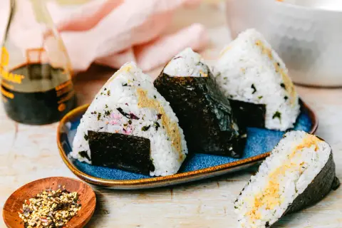 Drei dreieckige Onigiri mit Thunfisch-Mayo-Füllung, umhüllt von Nori-Algen, auf einem blauen Teller. Daneben eine Flasche Sojasauce und eine Schale mit Gewürzen.
