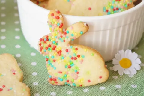 Bunte Osterkekse zum Ausstechen in Hasenform, verziert mit bunten Streuseln, liegen auf einem grünen Tupfentuch neben einer weißen Schüssel.
