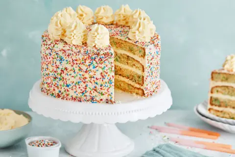 Eine leckere Torte mit weißem Zuckerguss und bunten Streuseln, gekrönt mit Sahnetuffs. Ein Stück ist angeschnitten und zeigt die saftigen Schichten.