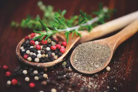 Zwei Holzlöffel mit Pfefferkörnern und gemahlenem Pfeffer. Ein Zweig Rosmarin liegt darüber. Ist Pfeffer gesund? Ja, er würzt und ist gut für dich!
