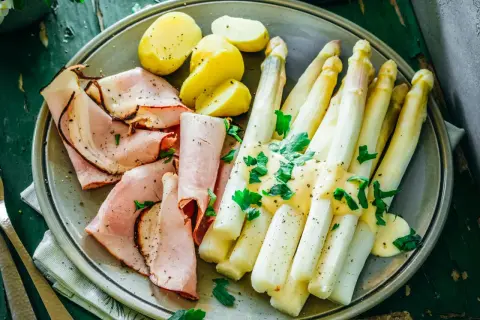 Ein Teller mit gekochtem Spargel, Schinken und Kartoffeln, garniert mit Petersilie. Perfekt für die besten Spargelrezepte!
