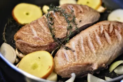 Zwei gebratene Entenbrüste mit Rosmarin, Apfelscheiben und Zwiebeln in einer Pfanne. Ein köstliches entenfilet rezept.