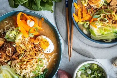 Zwei Schalen mit dampfendem, **vegetarischem Ramen**. Nudeln, Karottenstreifen, Pak Choi, Tofuwürfel und ein halbes Ei in herzhafter Brühe.