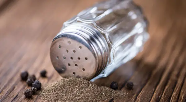 Ein Glasstreuer mit Metallaufsatz liegt auf einem Holztisch. Gemahlener Pfeffer und ganze Körner sind daneben verstreut. So kann man den Pfeffer neutralisieren.
