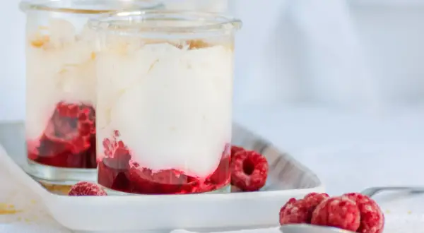 Zwei Gläser mit cremigem Dessert und Himbeeren. Unten leuchtet ein intensiver Himbeercoulis, gekrönt von luftiger Sahne. Frische Himbeeren zieren die Gläser und eine Löffelspitze.