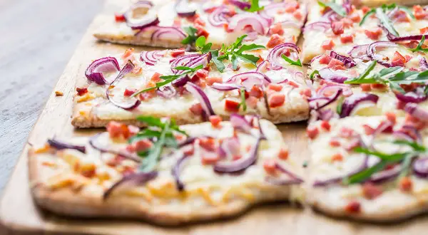 Flammkuchen ohne Speck, belegt mit roten Zwiebeln, Käse und Rucola, auf einem Holzbrett.