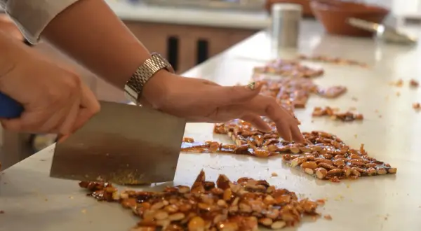 Eine Hand schneidet mit einem Messer karamellisierte Mandeln. So entsteht Nougat, das hauptsächlich aus Nüssen und Zucker besteht.