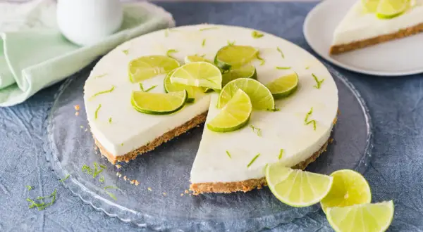 Erfrischender limetten cheesecake ohne backen, dekoriert mit frischen Limettenspalten und Zesten. Ein Stück ist bereits angeschnitten.