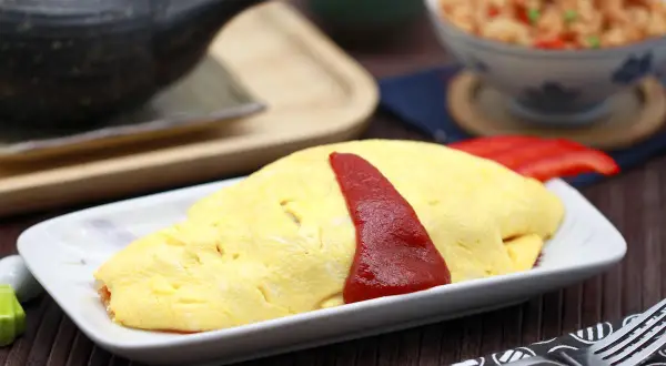 Ein leckeres Omurice Rezept: goldgelbes Omelett mit Ketchup-Streifen auf einem Teller, daneben eine Schüssel mit gebratenem Reis.