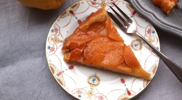 Ein Stück Quitten Tarte Tatin auf einem Teller mit Blumenmuster. Daneben eine ganze Quitte und ein weiteres Stück Tarte.