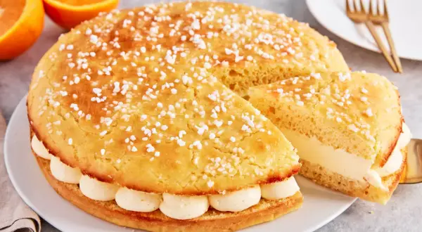 Eine köstliche Tarte Tropezienne, bestreut mit Zuckerperlen, gefüllt mit cremiger Vanillecreme. Daneben liegen Orangen.