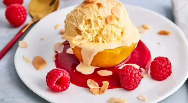 Ein Pfirsich Melba Rezept Original: Ein Pfirsich mit Vanilleeis, Himbeersauce und Mandelsplittern auf einem Teller, garniert mit frischen Himbeeren.