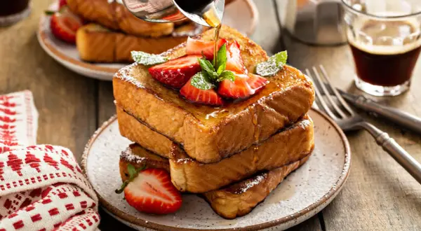 Ein Stapel goldbrauner Pain Perdu, garniert mit Erdbeeren und Minze, wird mit Sirup übergossen. Daneben ein Glas Kaffee.