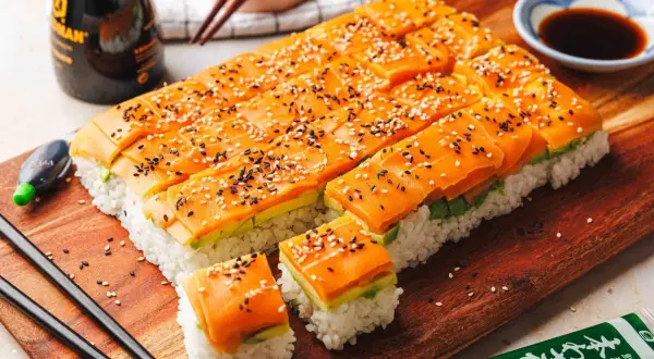 Leckere Sushi-Ideen: Reis, Avocado und Lachs, bestreut mit Sesam. Serviert auf einem Holzbrett mit Sojasauce und Wasabi.