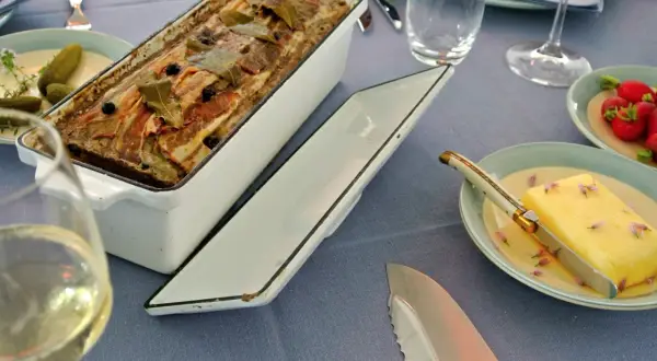 Fertige Terrine mit Speck und Lorbeerblättern, bereit für ein köstliches Terrinen Rezept. Dazu gibt es Butter, Radieschen und ein Glas Weißwein.