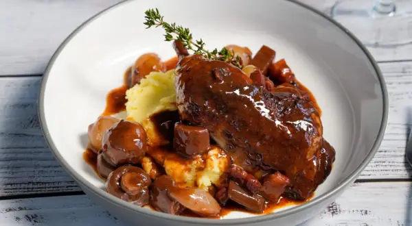 Ein authentisches Coq au Vin Rezept Original: zartes Hähnchen in Rotwein, serviert mit Kartoffelpüree, Speck und glasierten Zwiebeln.