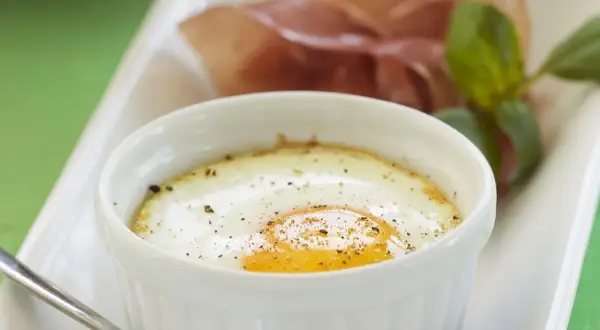 Ein köstliches oeuf cocotte mit einem perfekt gebratenen Ei, bestreut mit Pfeffer, serviert mit Schinken und Basilikum.
