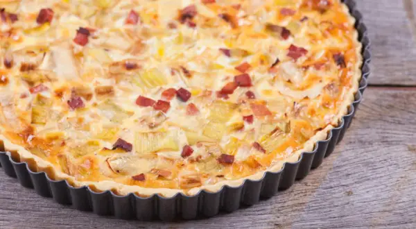 Eine herzhafte Quiche Lorraine mit Lauch und Speck in einer Metallform auf Holzhintergrund.