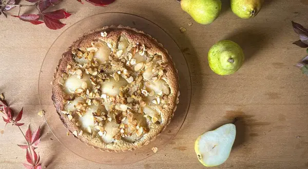 Eine köstliche Tarte Bourdaloue mit Birnen und Mandelsplittern, umgeben von frischen Birnen und herbstlichen Blättern auf Holzhintergrund.