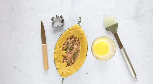 Ein ovaler Teigmantel, gefüllt mit Hackfleisch und Kräutern, wird mit Blätter-Ausstechern verziert. Daneben liegen ein Messer, ein Ausstecher, Eigelb und ein Pinsel für das perfekte Pate en croute.