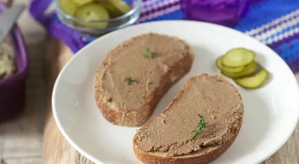 Zwei Scheiben Brot mit selbstgemachter Leberwurst, garniert mit Kräutern, dazu Gewürzgurken und ein lila Glas.