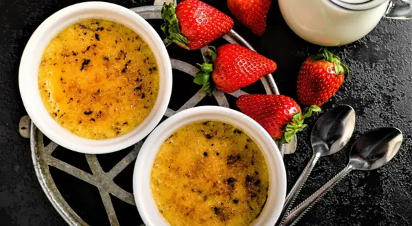 Zwei cremige Crème brûlée mit karamellisierter Zuckerkruste, garniert mit frischen Erdbeeren und einem Milchkännchen.